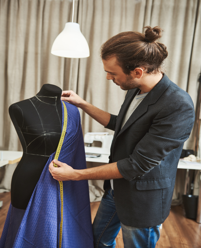 Vertical portrait of adult good-looking caucasian male fashion designer with stylish hairstyle in fashionable outfit in his studio working on new dress for winter clothes collection